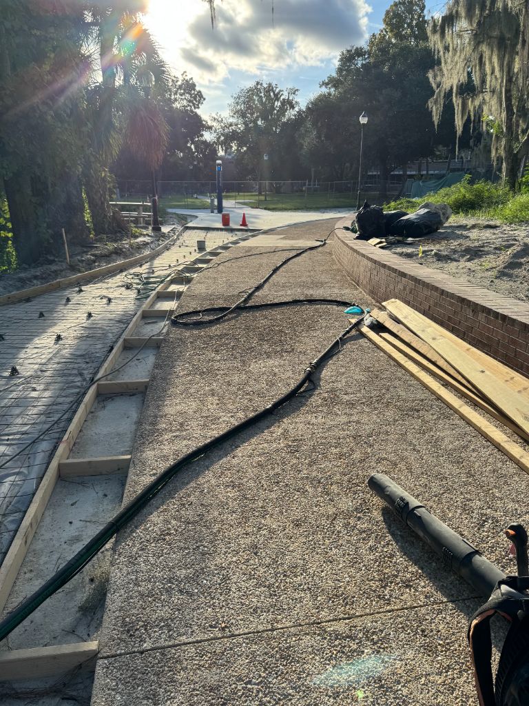 University of Florida campus sidewalk during sandblasting — blasting hoses deployed, top concrete layer being removed to reveal pebble aggregate surface, curved brick retaining wall visible